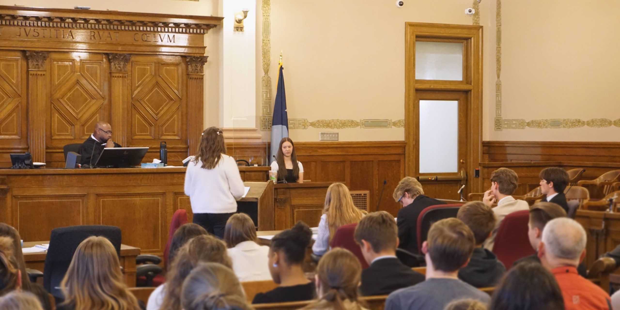 Attorney and witness interacting in a courtroom with a galley full of observers.