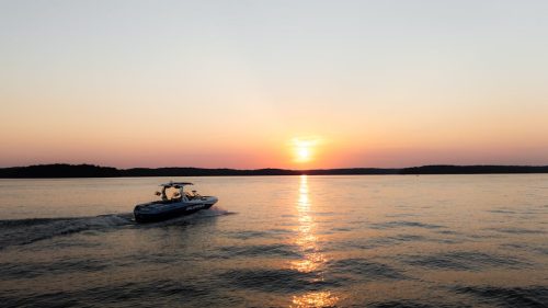 A speedboat navigates a serene lake as the sun sets, casting colorful reflections on the water.