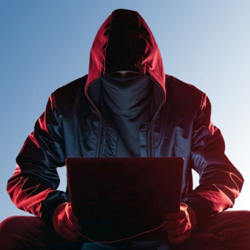 A person wearing a red hooded jacket and sitting cross-legged is using a laptop in a dark setting.